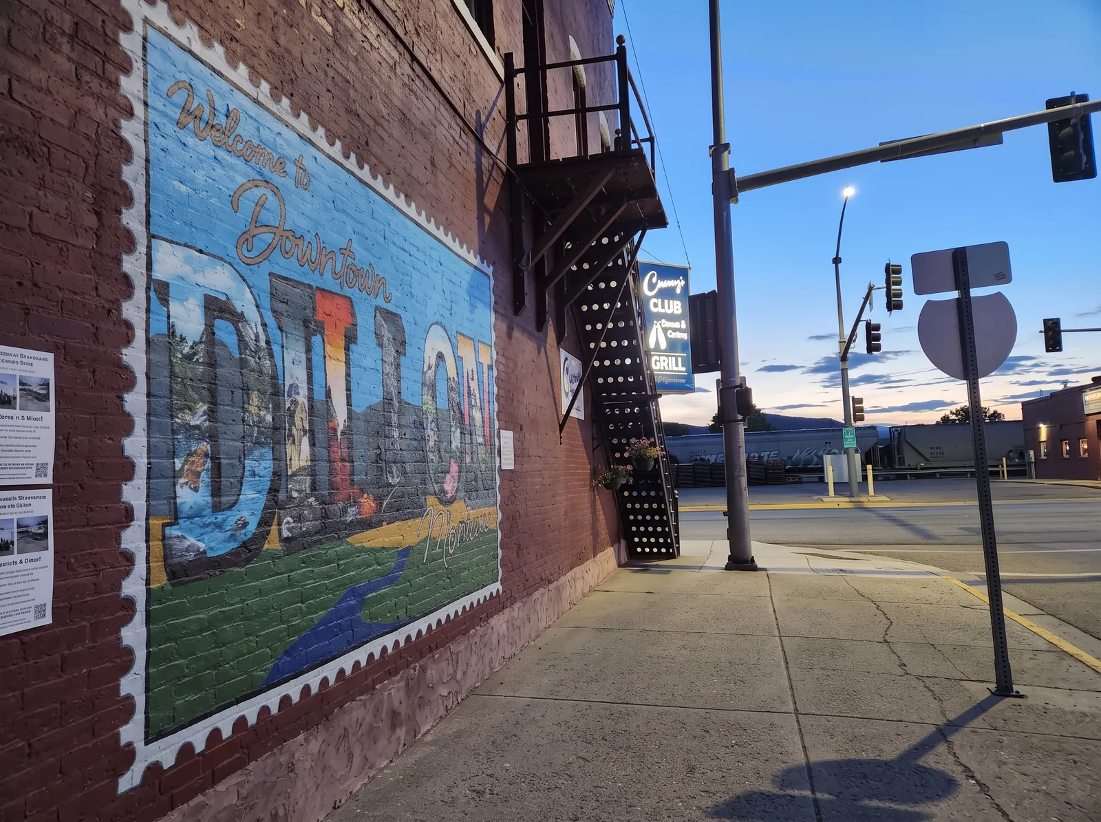 The Welcome to Downtown Dillon mural on the side of the Cranny's building at dusk, with the Club sign lit
